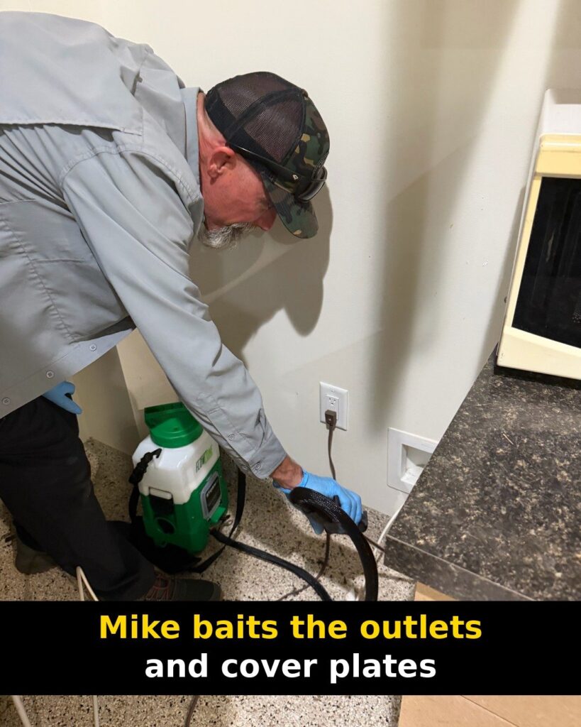 Pest control technician bending over near a green sprayer on the floor and a wall outlet in a break room